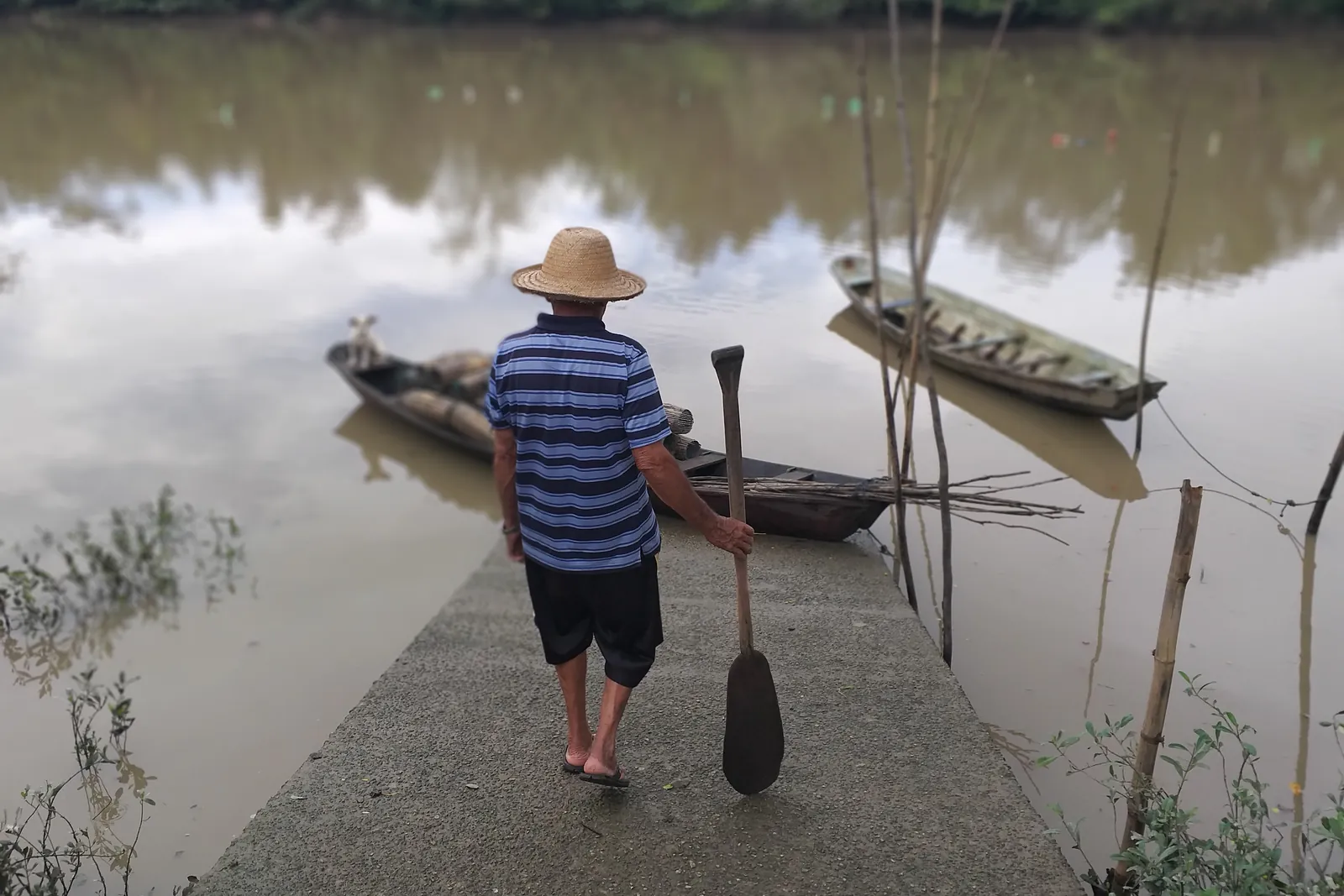 Pescador artesanal de chapéu de palha e camisa listrada, visto de costas, segurando um remo num trapiche diante de canoas no rio.